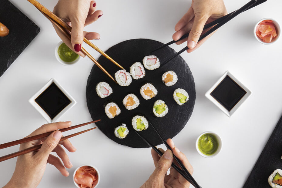 Manos agarrando sushi con palillos. Plato de sushi compartido con salsas y jengibre.
